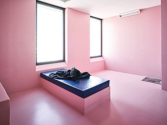 Das Bild zeigt eine Sicherheitszelle im Regionalgefängnis in Burgdorf mit zwei großen Fenstern. Die Wände sowie der Boden sind komplett in Rosa gestrichen. Im Raum befindet sich nur ein rosafarbenes Bett mit einer blauen Matratze.