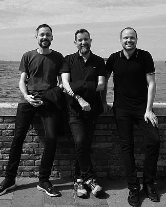  Three men are standing in front of a wall and in the background is the sea