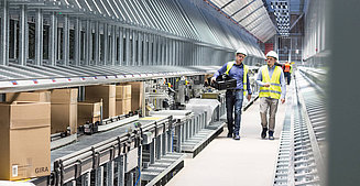  Two employees go through a hall in logistics, production and development takes place.