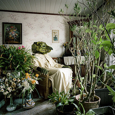 Eine als grünes Monster verkleidete Person sitzt in einem Wohnzimmer, umgeben von Pflanzen.