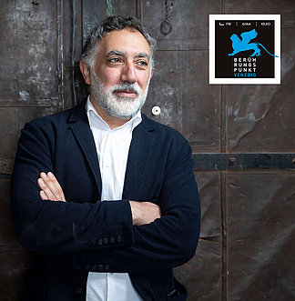 Portrait von Hashim Sarkis, Kurator der Architekturbiennale 2020 in Venedig vor einer Wand. Er verschränkt die Arme und trägt ein weißes Hemd und ein Blaues Jackett. 